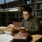 A young man in a sweater reads books in a library, surrounded by books and a laptop.